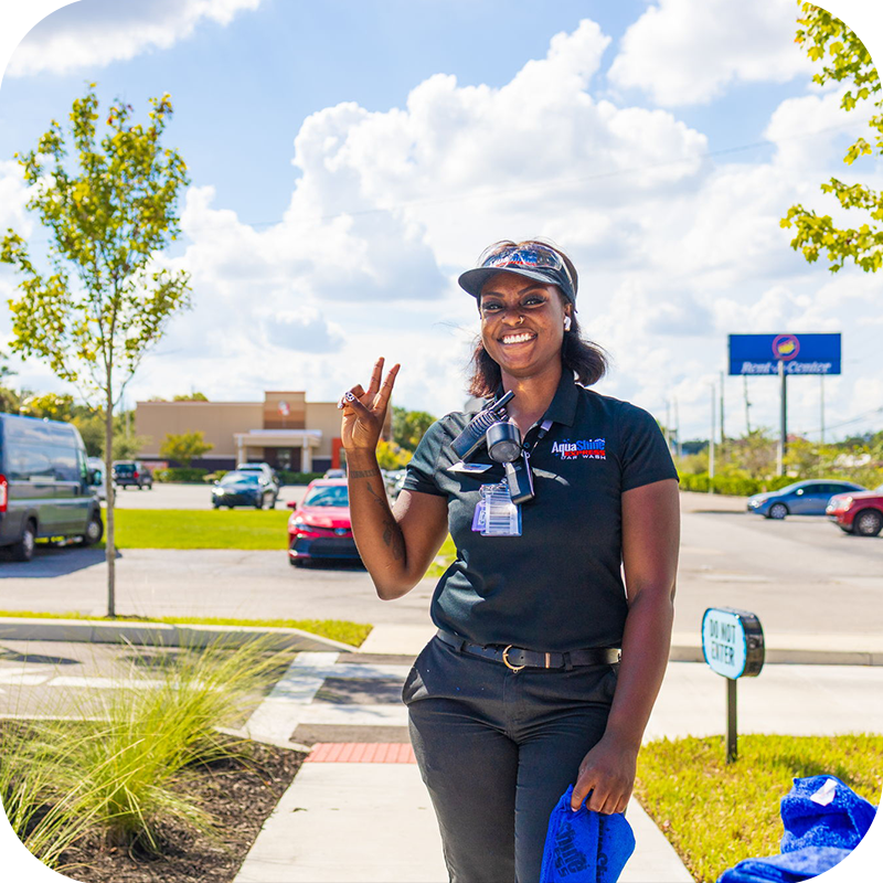 Smiling AquaShine Express Car Wash team member standing outside the car wash and waving at customers